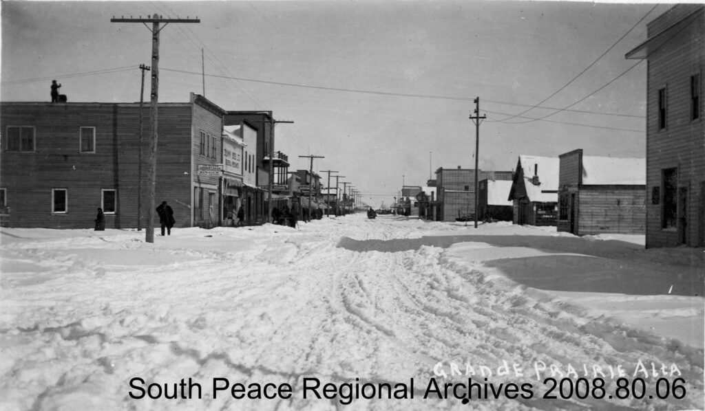 Grande Prairie's main street showing businesses lining both sides and a hose and wagon disappearing down a rutted snow path. (ca. 1930)